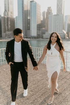 a man and woman holding hands while walking by the water in front of city buildings