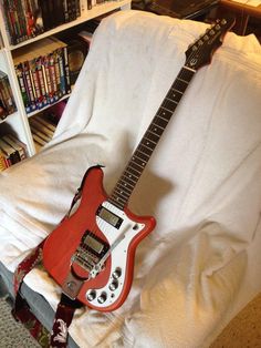 a red electric guitar sitting on top of a white couch next to a book shelf