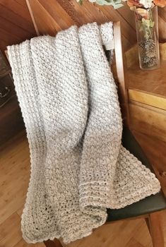 a white knitted blanket sitting on top of a wooden chair next to a vase with flowers