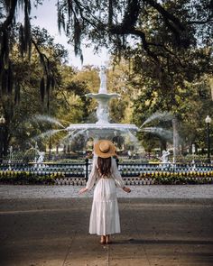 a woman in a white dress and straw hat standing in front of a fountain