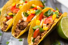 three tacos with meat, corn and tomatoes on a wooden table next to a lime