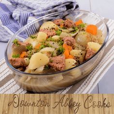 a glass bowl filled with meat and vegetables on top of a wooden table next to a towel