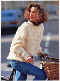 a woman riding on the back of a bike with a basket in her hand while wearing a sweater and jeans