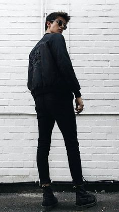 a man standing in front of a white brick wall wearing sunglasses and a black jacket