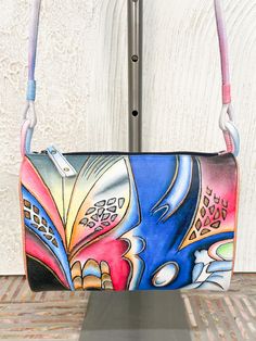 This artful cross-body style has classic satchel shape, with a top zip closure and outer zip closure at back side. Interior features credit card slot organization. Non-adjustable crossbody strap is attached with two leather rings at sides. Hand-painted Genuine leather 9.5” wide x 7.5” high x 3” deep Made in India Style # 1781 Artistic Leather Shoulder Bag With Adjustable Strap, Artistic Blue Shoulder Bag For Everyday Use, Artistic Hand Painted Crossbody Shoulder Bag, Artistic Blue Shoulder Bag, Artistic Leather Bags With Adjustable Strap, Artistic Crossbody Bag With Adjustable Strap, Artistic Crossbody Shoulder Bag For Travel, Hand Painted Multicolor Shoulder Bag For Everyday Use, Hand Painted Crossbody Travel Bag