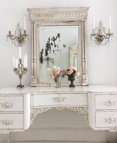 a white desk with flowers and candles on it in front of a mirror, chandelier and wall sconces