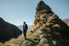 a man in a suit standing on top of a grass covered hill next to a rock formation