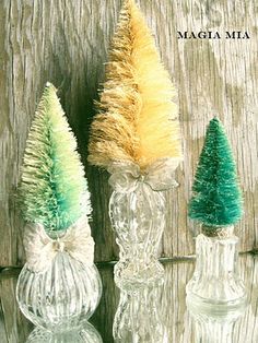 three glass vases with fake trees in them on a wooden surface, one is green and the other is yellow