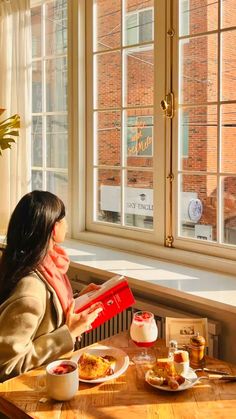 a woman sitting at a table in front of a window reading a book and eating