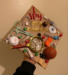 a person's hand holding up a graduation cap with school supplies on it and the words i spy