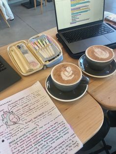 two cups of cappuccino sit on a table next to an open notebook