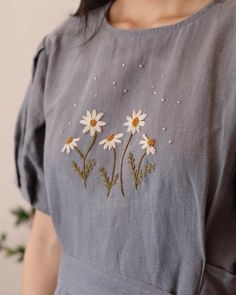 a woman wearing a gray dress with daisies embroidered on the chest and shoulder, standing in front of a white wall