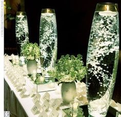 two tall vases filled with green plants on top of a white cloth covered table