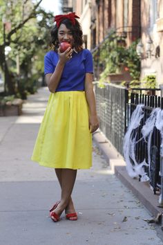 a woman in a yellow skirt eating an apple while standing on the sidewalk next to a fence