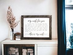 a wooden framed sign sitting on top of a white shelf next to a window and vase with flowers