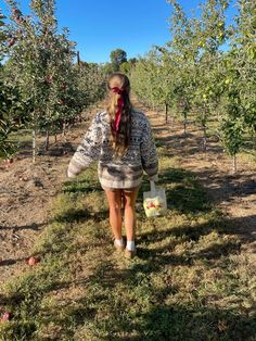 a woman walking through an apple orchard carrying a bag