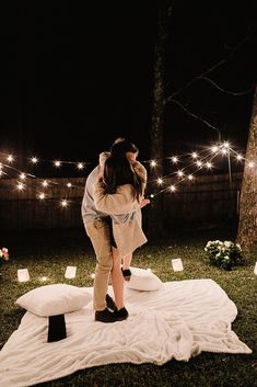 a man and woman kissing on a blanket in the grass with lights strung above them