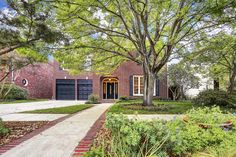 a brick house surrounded by trees and flowers in the front yard with a pathway leading to it