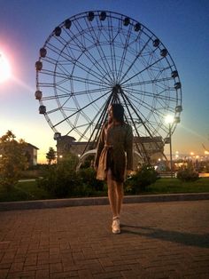 a woman is standing in front of a large ferris wheel at night with the sun setting behind her