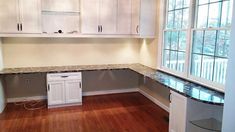 an empty kitchen with white cabinets and black counter tops on hard wood flooring in front of large windows