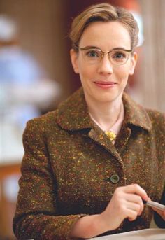 a woman with glasses is holding a piece of paper and looking at the camera while sitting down