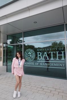 a woman standing in front of a bath school entrance wearing a pink suit and white sneakers