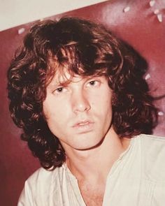 a man with long curly hair wearing a white shirt and looking at the camera while standing in front of a red wall