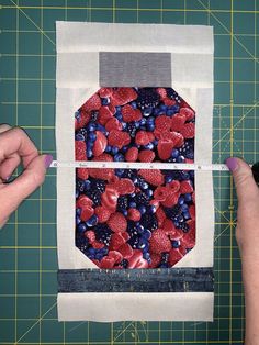 two hands are holding a piece of fabric with berries and blueberries on it, while another hand is measuring the length