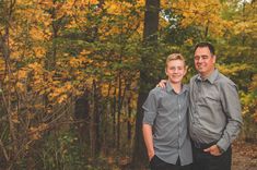 two men standing next to each other in front of trees with leaves on the ground