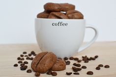 coffee beans spilling out of a white cup on top of a wooden table next to a pile of chocolate cookies