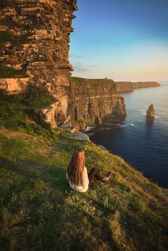 a woman sitting on top of a lush green hillside next to the ocean with cliffs in the background