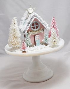 a white cake plate topped with a small pink house and trees covered in frosting