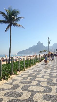 people are walking on the sidewalk near the beach and palm trees in front of some mountains