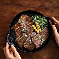 two hands holding a skillet with steak and vegetables on it, while another person holds the pan