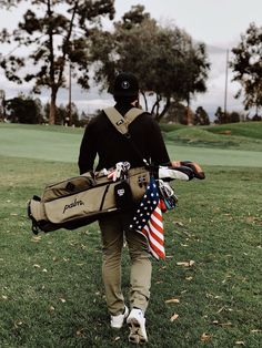 a man walking across a lush green field holding a golf bag and an american flag