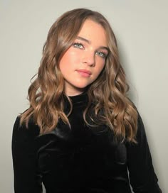 a woman with long hair wearing a black dress and posing in front of a gray wall