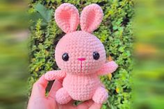 a small pink crocheted bunny sitting on top of a lush green field with bushes in the background