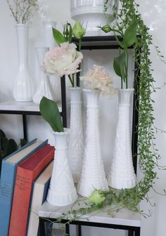 four white vases with flowers and greenery in them on a shelf next to books