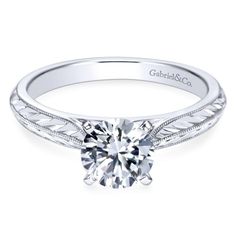 a white gold engagement ring with an intricate design on the band and a round diamond center stone