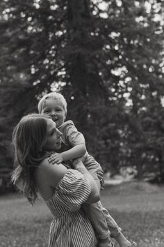 a woman holding a small child in her arms while standing on the grass with trees in the background