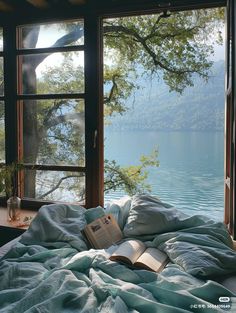 an open book sitting on top of a bed in front of a window next to a lake