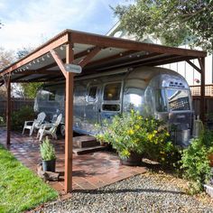 an airstream is parked in the yard under a pergolated awning with two chairs