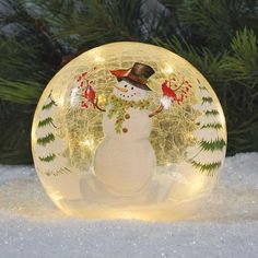 a glass snowman ornament sitting in the snow next to a pine tree