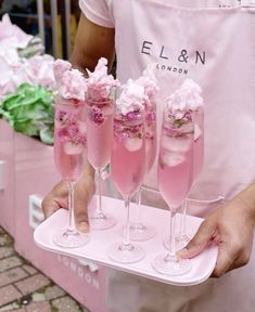 a person holding a tray with five glasses filled with pink liquid and topped with flowers