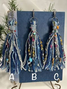 two blue earrings with beads and chains hanging from hooks on a metal stand next to a pine tree