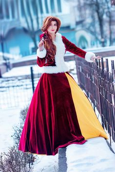 a woman in a long red and yellow dress standing next to a fence with snow on it