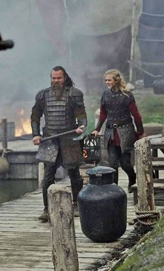 two people dressed in medieval clothing standing on a dock next to a large black pot