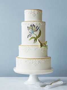 a three tiered wedding cake decorated with flowers and butterflies on a white pedestal next to a blue wall