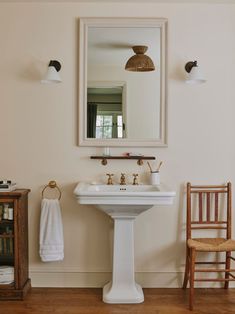 a white pedestal sink sitting under a bathroom mirror next to a wooden chair and table