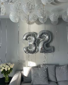 silver balloons are hanging from the ceiling in front of a couch and table with white flowers